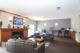This single-floor space is thoughtfully designed to offer multiple distinct areas for gathering, relaxing, and socializing. The entry foyer sets a warm tone with natural wood-framed glass doors, tile flooring, and a small seating arrangement of dark leather chairs around a round table, surrounded by lush indoor plants and views of the outdoors through a large sliding window.

The main lounge area centers around a striking wood-trimmed fireplace with a slate surround, flanked by windows that bring in natural light. A wall-mounted flat-screen TV sits above the mantel, which is dressed with decorative accents. Dark leather sofas are arranged around a rustic wood coffee table atop a vibrant, multicolored area rug, creating an inviting focal point.

A secondary lounge offers a more subdued atmosphere, featuring a slate-blue accent wall with sconce lighting, leather seating with blue accent pillows, and a built-in counter with bar-height seating—ideal for working or casual use.

The kitchen is well-appointed with warm wood cabinetry, dark countertops, black appliances, a tile backsplash, and a spacious island with bar stool seating. Pendant lighting and a mounted TV complete the functional yet welcoming space.