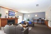 This single-floor space is thoughtfully designed to offer multiple distinct areas for gathering, relaxing, and socializing. The entry foyer sets a warm tone with natural wood-framed glass doors, tile flooring, and a small seating arrangement of dark leather chairs around a round table, surrounded by lush indoor plants and views of the outdoors through a large sliding window.

The main lounge area centers around a striking wood-trimmed fireplace with a slate surround, flanked by windows that bring in natural light. A wall-mounted flat-screen TV sits above the mantel, which is dressed with decorative accents. Dark leather sofas are arranged around a rustic wood coffee table atop a vibrant, multicolored area rug, creating an inviting focal point.

A secondary lounge offers a more subdued atmosphere, featuring a slate-blue accent wall with sconce lighting, leather seating with blue accent pillows, and a built-in counter with bar-height seating—ideal for working or casual use.

The kitchen is well-appointed with warm wood cabinetry, dark countertops, black appliances, a tile backsplash, and a spacious island with bar stool seating. Pendant lighting and a mounted TV complete the functional yet welcoming space.