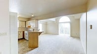 This single-floor space features an open-concept layout that seamlessly connects the kitchen and living area, creating a cohesive and functional environment. The kitchen is equipped with light wood cabinetry, a gas range, and a peninsula-style breakfast bar with warm oak detailing, providing both additional counter space and a casual dining option. Tile flooring grounds the kitchen area, transitioning into plush wall-to-wall carpeting that extends throughout the spacious living area.

The living space is bright and airy, benefiting from multiple sources of natural light. A prominent arched window adds an architectural focal point, while large sliding glass doors open to an outdoor balcony, further enhancing the sense of openness and allowing views of the surrounding exterior. Vertical blinds on the sliding doors provide privacy and light control as needed.

A structural beam subtly defines the boundary between the kitchen and living zones without interrupting the open flow of the space. The neutral color palette throughout — from the cream-toned walls to the light carpet — creates a clean, versatile backdrop well-suited to a variety of interior styles.