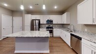 This single-floor residence offers a thoughtfully designed layout that balances open communal spaces with comfortable private quarters. The kitchen serves as a centerpiece of the home, showcasing crisp white shaker-style cabinetry, granite countertops with a subtle grey-and-white veining pattern, and a full suite of stainless steel appliances including a refrigerator, electric range, over-the-range microwave, and dishwasher. A large kitchen island provides additional counter space and seating, while pendant lighting and recessed fixtures illuminate the area effectively. The kitchen flows seamlessly into the living area, which features warm hardwood-style flooring, neutral grey walls, recessed lighting, a ceiling fan, and a door leading to an outdoor space with natural light filtering through the windows.

The bedrooms are carpeted for comfort and each includes a ceiling fan, a window for natural light, and ample space for furnishings. At least one bedroom features a walk-in closet with built-in shelving for organized storage. The bathrooms are clean and functional, with one featuring a full tub-shower combination, wood-style flooring, and a vanity with modern hardware. The consistent use of neutral tones, white trim, and wood-style flooring throughout creates a cohesive and contemporary aesthetic across the entire residence.