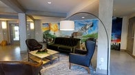 This single-floor space makes a strong first impression with its thoughtfully curated design and artistic character. The lounge area serves as the centerpiece, anchored by a dramatic large-scale mural that spans the wall, depicting a vivid cityscape with colorful, painterly strokes — instantly establishing the space's creative identity.

The furnishings strike a balance between sophistication and comfort. A tufted dark green velvet sofa commands attention at the center of the room, flanked by sleek black leather armchairs that offer both style and functionality. A glass-and-metal coffee table adds an industrial edge, complementing the overall aesthetic. An oversized arc floor lamp with a white drum shade provides a warm, focused glow, enhancing the lounge's intimate yet open feel.

The walls feature a smooth concrete-style finish in cool gray tones, contributing to the modern-industrial character of the space. Recessed ceiling lighting works in harmony with the arc lamp to create layered illumination throughout. A woven area rug in muted tones anchors the seating arrangement, adding texture and warmth underfoot.

Overall, the space exudes a curated, urban-chic ambiance — ideal for socializing or relaxing — blending artistic expression with contemporary design elements seamlessly.