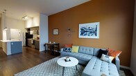 This single-floor space presents a thoughtfully designed open-concept layout that combines the living, dining, and kitchen areas into one cohesive environment. The living area serves as the focal point, anchored by a bold terracotta-orange accent wall that adds warmth and character to the space. A contemporary light-grey sectional sofa, adorned with colorful throw pillows, is paired with a sleek round white coffee table and a patterned area rug, creating an inviting and modern seating arrangement. Decorative wall art, including a large landscape photograph and starburst accents, add personality to the space. The dining area transitions naturally from the living room, featuring a compact wooden table with matching chairs — an efficient use of space that maintains an open, airy feel. The kitchen is visible just beyond, equipped with stainless steel appliances including a range and over-the-range microwave, set against clean cabinetry and countertops. Dark hardwood flooring runs throughout, providing a rich contrast to the lighter furnishings and wall tones. The overall ambiance is modern and welcoming, with a curated mix of textures and colors that make the space feel both functional and stylish.