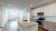 This single-floor residence showcases a thoughtfully designed open-concept layout where the kitchen flows seamlessly into the living area. The kitchen is a standout feature, offering a large central island with granite countertops and an integrated sink, complemented by a two-tone cabinetry scheme pairing white upper shaker-style cabinets with warm gray lower cabinets. A classic white subway tile backsplash frames the full suite of stainless steel appliances, including a French door refrigerator, electric range with over-the-range microwave, and recessed lighting throughout. Rich, dark hardwood-style flooring extends across the kitchen and living spaces, adding warmth and visual continuity. The spacious living area benefits from natural light provided by multiple windows with horizontal blinds and is equipped with a ceiling fan. A separate bedroom features plush carpet flooring and generous wall space, while the bathroom includes a granite-topped vanity with taupe cabinetry, a tub-shower combination, wood-style flooring, and direct access to a walk-in closet with built-in shelving. Clean white walls and recessed lighting throughout create a bright, modern ambiance.
