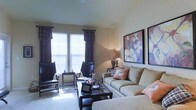 This single-floor space offers a comfortable and thoughtfully designed living environment. The living room serves as the centerpiece, showcasing a warm, neutral color palette with beige walls that create an inviting atmosphere. Large windows dressed with dark decorative curtains and horizontal blinds allow abundant natural light to fill the room, casting a bright and airy feel throughout.

The living room is generously sized, accommodating a plush, neutral-toned sofa accented with decorative throw pillows in soft coral and patterned hues. A sleek glass-top coffee table anchors the seating arrangement, complemented by a pair of dark leather accent chairs and a coordinating side table. The combination of furnishings creates a balanced and functional layout ideal for both relaxing and entertaining.

The walls are adorned with bold, large-format botanical artwork in warm earth tones, adding a tasteful decorative focal point to the space. Soft carpeted flooring contributes to the room's cozy ambiance. A floor lamp provides additional ambient lighting, enhancing the welcoming atmosphere. The overall design reflects a harmonious blend of comfort and style, making this a well-rounded and livable space.