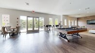 This single-floor amenity space is thoughtfully designed to offer a range of social and recreational experiences within a cohesive, modern aesthetic. A recurring design element throughout is the reclaimed wood plank wall paneling, rendered in soft gray and taupe tones, which lends warmth and a contemporary rustic character to the interior.

The game room is a spacious, open area with rich hardwood-style flooring, a full-size billiards table, and bar-height seating. Large glass doors open to an outdoor terrace, flooding the space with natural light and providing views of surrounding greenery.

Adjacent to the game room, a lounge area features the same signature wood-paneled walls, now serving as a backdrop for a large flat-screen television mounted at eye level. Comfortable modern seating and patterned carpeting create an inviting setting for casual gatherings.

A dedicated relaxation nook offers an intimate retreat, featuring two tufted chaise lounges with decorative pillows and soft ambient lighting—ideal for unwinding.

The café-style dining area showcases pendant lighting, teal upholstered banquette seating, and pedestal tables paired with Scandinavian-inspired chairs. Nearby, a well-appointed kitchenette provides dark cabinetry, stainless steel appliances, and ample counter space to support entertaining and everyday use.
