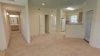 This single-floor residence offers a clean, open layout that seamlessly connects the main living area to the kitchen, creating a spacious and functional flow throughout. The living space is finished with plush wall-to-wall carpeting in a warm neutral tone, contributing to a calm and comfortable ambiance. Soft, cream-colored walls are found throughout, reflecting light and enhancing the sense of openness.

The kitchen is positioned at the edge of the main living area and features white cabinetry, a full-size refrigerator, and counter space with an arched window above the sink that allows natural light to pour in. A raised countertop serves as a natural divider between the kitchen and living space.

Recessed lighting and a fluorescent overhead fixture provide ample illumination across the open-plan area, while visible HVAC vents ensure year-round climate control. A doorway from the main area leads to an adjacent room, also carpeted and well-lit by natural light, suggesting additional living or sleeping space. A closet with a paneled door is accessible nearby, providing convenient storage.

Overall, the space presents a bright, neutral palette with a practical open-concept design suited for comfortable everyday living.