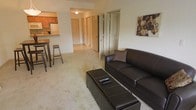 This single-floor residence offers an open and functional layout that seamlessly connects the main living area to the kitchen. The living room is carpeted in a neutral, light-toned material that contributes to a warm and comfortable atmosphere. It is furnished with a dark leather sofa accented by a decorative pillow, paired with a matching ottoman-style coffee table that doubles as additional seating or storage. Decorative wall art adds a subtle focal point to the space.

Adjacent to the living room, a raised bar counter with tall stools creates a casual dining area that opens directly to the kitchen. The kitchen features warm wood-toned cabinetry and includes standard appliances such as a refrigerator. The space is illuminated by overhead lighting, maintaining a bright and practical environment.

A hallway visible from the living area leads to multiple interior doors, indicating access to bedrooms, closets, or additional rooms. The overall layout is straightforward and efficient, making good use of the available floor space. The neutral color palette throughout — from the walls to the flooring — creates a cohesive, understated aesthetic well-suited for everyday living.