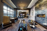 Located at 119 Searing Avenue in Mineola, New York, this upscale amenity lounge is a thoughtfully designed communal space that blends contemporary style with comfortable functionality. The single-floor layout encompasses several distinct zones that flow seamlessly into one another, unified by designer patterned carpeting and a refined dark-and-neutral color palette throughout.

The lounge area serves as the centerpiece, anchored by a dramatic floor-to-ceiling iridescent brick accent wall housing a sleek linear gas fireplace. High-back wingback chairs with bold geometric upholstery are arranged to create an intimate seating environment. Nearby, oversized floor-to-ceiling windows flood the space with natural light while offering open views of the surrounding streetscape, with sheer curtains softening the light during peak hours.

A stylish bar area features a white countertop with decorative tile detailing, accompanied by tall bar stools and wall-mounted flat-screen televisions — ideal for social gatherings. Adjacent to the bar, a billiards table sits beneath a sculptural pendant light fixture, adding a recreational focal point to the space.

Throughout, coffered ceilings with recessed lighting and statement chandeliers add architectural depth. A vestibule entry with glass-paneled French doors and curated artwork provides an elegant transition into the main lounge, completing this well-appointed amenity space.