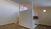 This single-floor residence offers a straightforward and functional layout designed for comfortable everyday living. The space is defined by neutral cream-toned walls and light-colored ceilings that create a bright, cohesive atmosphere throughout. A distinctive half-wall partition serves as an architectural divider, subtly separating the living area from the kitchen while maintaining an open, connected feel. A simple pendant light fixture hangs above, adding a understated focal point to the shared space. The kitchen features white cabinetry, a dark countertop, and tile flooring, providing a clean and practical workspace. White appliances and a baseboard heating unit are also visible, reflecting a no-frills, utilitarian design approach. The living area is carpeted, adding a layer of warmth underfoot and contrasting with the tile in the kitchen zone. Toward the rear of the space, a walk-in closet area with shelving and hanging rods offers organized storage. The overall ambiance is calm and minimal, with an emphasis on practicality and ease of use. The layout flows naturally from one area to the next, making efficient use of the available footprint across the single level.