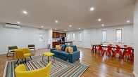 Located at 587 Broadway in Menands, NY, this single-floor community amenity space offers a thoughtfully designed layout that balances fitness, socializing, and relaxation. Warm hardwood-style flooring runs throughout, tying the various areas together with a cohesive, inviting feel.

The entrance hallway provides a clean, bright welcome point, featuring a console table and frosted-glass door leading into the gym. The exercise room is generously sized, outfitted with multiple treadmills and stationary bikes, rubber flooring, bright overhead panel lighting, and large windows that bring in natural light and views of the surrounding brick architecture.

The open-concept living, dining, and kitchen area is the heart of the space. The kitchen is well-equipped with rich wood cabinetry, granite countertops, stainless steel appliances including a range, microwave, and refrigerator, and a white subway tile backsplash. A row of tall windows fills the area with natural light. Adjacent to the kitchen, a spacious dining table with vibrant orange metal chairs seats a crowd comfortably. The living area features a plush blue sofa, colorful accent chairs, and a patterned area rug that adds personality.

A separate living room offers additional seating with yellow accent chairs and a wall-mounted climate control unit, completing this versatile and energetic communal space.