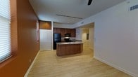 This single-floor space showcases an open-concept layout that seamlessly connects the kitchen and living areas. The kitchen serves as the centerpiece, featuring rich medium-toned wood cabinetry that extends to the upper walls, providing ample storage. A central kitchen island with a built-in sink and neutral countertop offers both prep space and a natural gathering point. Black appliances, including a refrigerator, microwave, and range, complement the warm cabinetry tones. Track lighting runs along the kitchen ceiling, supplemented by pendant lights above the island, ensuring well-distributed task lighting. Recessed lighting is also integrated throughout the space for ambient illumination. Light-toned hardwood flooring extends across the entire area, lending warmth and visual continuity. A set of bi-fold closet doors along one wall adds practical storage while maintaining the clean aesthetic. Neutral white walls contrast pleasantly with the darker wood tones. A hallway visible in the background connects to additional areas of the unit, suggesting a functional and well-organized floor plan overall.