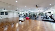 Located at Harbor Pointe in Sandy Springs, GA, this fitness center offers a generously sized, single-floor exercise facility designed to support a wide range of workout routines. The space is divided into two distinct functional zones: a hardwood-floored open area ideal for stretching, yoga, or group exercise, and a rubber-floored equipment zone housing an extensive selection of strength and cardio machines. The strength training section includes cable machines, weight racks, a full dumbbell rack, adjustable benches, and stability balls, while the cardio area features treadmills, stationary bikes, and recumbent machines. Floor-to-ceiling mirrors line the walls, visually expanding the space and allowing users to monitor their form throughout their workouts. Large windows along one side of the room invite abundant natural light and offer views of the surrounding greenery, creating a bright and energizing atmosphere. Multiple ceiling fans are distributed throughout the space, ensuring consistent airflow and comfort. Recessed lighting provides even illumination throughout all areas. Additional private rooms or utility spaces are accessible along one corridor, complemented by seating nooks for rest between sets. The overall design is clean, functional, and well-maintained, making this fitness center a comprehensive resource for residents.