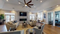 Located at 2700 Whakatane Lane in Fuquay-Varina, NC, this single-story community clubhouse offers a thoughtfully designed layout with distinct areas for relaxation, productivity, and recreation. The main lounge area is the centerpiece of the space, featuring soaring vaulted ceilings, recessed lighting, and a large ceiling fan that enhance the open, airy atmosphere. A floor-to-ceiling stone-accented fireplace wall anchors the room, complete with a mounted flat-screen television, while multiple seating arrangements on patterned area rugs invite gathering and conversation. French doors provide direct outdoor access and flood the space with natural light, complemented by large windows throughout. Rich hardwood-style flooring flows consistently across the entire facility. The kitchen area features white shaker cabinetry, granite countertops, a blue tile backsplash, stainless steel appliances, and a coffee station. A casual dining nook with a high-top table and leather barstools offers a spot for informal gatherings, benefiting from views of the surrounding landscape. The business center provides multiple workstations with built-in white cabinetry, desktop computers, and botanical-themed artwork. A game room accommodates a pool table and shuffleboard, while a fitness area rounds out the amenities, making this clubhouse a versatile hub for community life.