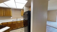 This single-story space includes a kitchen that combines practicality with a warm, welcoming aesthetic. The kitchen is equipped with honey-toned oak cabinetry that provides ample storage along the lower and upper sections of the room. A double-basin stainless steel sink is positioned beneath the cabinetry, offering a functional workspace. A full-size black refrigerator serves as a prominent feature, positioned at the entrance to the kitchen and contributing to a cohesive, modern appliance palette.

One of the most notable architectural features of the kitchen is the large skylight panel installed in the ceiling, which floods the space with bright, diffused natural light and creates an open, airy atmosphere. The light wood-toned flooring runs throughout, complementing the warm cabinetry tones and contributing to a cohesive interior palette. The walls are finished in a neutral beige tone, providing a clean backdrop that ties the space together. Overall, the kitchen presents a well-lit, organized environment suited for everyday use.