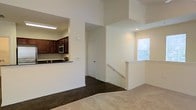 This single-floor space showcases an open and functional layout that connects the living area to the kitchen through a convenient pass-through opening. The kitchen features rich, dark wood cabinetry paired with dark countertops, stainless steel appliances including a built-in microwave, and recessed lighting that illuminates the workspace effectively. The pass-through counter creates a seamless visual connection between the kitchen and the main living area, enhancing the open feel of the space.

The living area is finished with wall-to-wall carpet in a neutral tone, contributing to a warm and comfortable atmosphere. Crisp white walls throughout create a clean, bright backdrop, while recessed lighting and a window fitted with blinds allow natural light to filter into the space.

A staircase with a white-painted railing is visible near the window, suggesting access to a lower level or entry area. A closed interior door offers additional privacy or storage. The overall design maintains a cohesive, neutral color palette, balancing practicality with a tidy, understated aesthetic throughout the space.