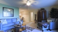 This single-floor residence offers a thoughtfully designed open-concept layout that seamlessly connects the living and dining areas, creating a spacious and welcoming atmosphere throughout. The living room is anchored by a ceiling fan with integrated lighting, providing both comfort and illumination. Soft, neutral-toned carpeting runs underfoot, complementing the warm beige and green accent walls that lend the space a calm, inviting character. A decorative area rug with a bold floral pattern adds visual interest beneath the centrally placed coffee table, while plush seating arrangements make the living area ideal for relaxing or entertaining. A large dark-wood armoire serves as a striking focal point along one wall, adding a rich, traditional element to the room's decor. The dining area, visible from the living space, is illuminated by an elegant chandelier and features classic dining furniture, reinforcing the cohesive and comfortable aesthetic of the home. A hallway leading off the main living area provides access to the remainder of the residence. The overall ambiance is warm, functional, and welcoming, with a harmonious blend of traditional furnishings and practical design elements that make this single-floor home both livable and visually appealing.