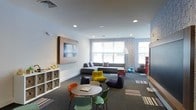 This single-floor space is a thoughtfully designed recreational lounge that balances comfort, functionality, and playful energy. The room features a clean, modern aesthetic with white ceilings equipped with recessed lighting, ensuring the space is well-lit throughout. Large windows along the far wall allow natural light to fill the room, creating a bright and welcoming atmosphere.

The lounge is organized into distinct activity zones. A cozy seating area anchors one side of the room, featuring a low-profile gray sectional sofa accented with colorful throw pillows, complemented by casual bean bag chairs in rich purple and yellow tones. A round activity table surrounded by vibrant multi-colored chairs provides an interactive space for games or casual gatherings.

A wall-mounted flat-screen display is prominently positioned on one wall, framed by a warm wood surround, making it a natural focal point. Open cube shelving nearby holds toys and accessories, adding a functional and organized touch.

The flooring transitions between dark carpet and patterned area rugs, adding texture and defining the various zones within the room. A striking textured stone-effect accent wall introduces depth and visual interest. Overall, the space exudes a lively, inviting ambiance well-suited for community activities, entertainment, and relaxation.