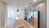 Located at 2220 Wilkinson Boulevard in Charlotte, NC, this thoughtfully designed one-bedroom, one-bathroom unit offers a modern aesthetic with high-quality finishes throughout. The open-concept living, dining, and kitchen area creates a seamless flow, anchored by a central kitchen island with a white quartz countertop and an undermount sink with a matte black faucet. The kitchen is equipped with stainless steel appliances — including a French door refrigerator, electric range, and built-in microwave — complemented by sleek gray cabinetry and a stone tile backsplash. Stylish pendant lighting adds a contemporary touch overhead. Light wood-look flooring extends throughout the living areas, enhancing the bright, airy ambiance created by recessed lighting and large windows. The living area opens to a private balcony offering open views of the surrounding landscape and Charlotte skyline. The bedroom is a comfortable retreat featuring a ceiling fan, access to a dedicated closet, and an adjacent bathroom. The bathroom is finished with large-format white tile on both the floor and walk-in shower walls, paired with matte black fixtures for a clean, modern look. Additional utility and laundry spaces round out this efficiently designed unit.