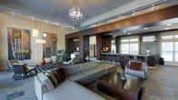 This single-floor space showcases a refined contemporary design with an open-concept layout that seamlessly connects multiple functional areas. The main lounge area is anchored by a striking crystal chandelier that serves as a dramatic focal point, complemented by a track lighting system that runs along the ceiling, providing versatile illumination throughout the space.

The seating area features large, neutral-toned sectional sofas adorned with richly colored accent pillows in deep greens and burgundies, creating an inviting yet sophisticated atmosphere. A dark wood coffee table grounds the arrangement, while bold abstract artwork decorates the walls, adding visual interest and personality to the space.

A defining architectural feature is the dark wood framing that elegantly separates the lounge from the adjoining kitchen and bar area. The kitchen and bar zone is equipped with a row of sleek bar stools lining a counter, recessed lighting, and modern cabinetry in rich, dark tones. Large windows in the bar area allow natural light to filter into the space. The overall ambiance is polished and upscale, blending warm wood tones with contemporary furnishings to create a welcoming and stylish environment.