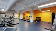 This fitness center offers a practical and energizing workout environment spread across a single level. The space is defined by its bold yellow accent walls, which add a vibrant, motivating atmosphere against the clean white structural columns and exposed ceiling infrastructure. The industrial-style ceiling features visible ductwork and fluorescent strip lighting, providing ample, even illumination throughout the area.

The flooring consists of durable dark grey tiles, well-suited for high-traffic exercise use. The cardio zone is equipped with multiple treadmills and a rowing machine, while a stationary bike occupies a separate area toward the back of the space, allowing for defined workout zones. A dedicated free weights section includes a multi-tiered dumbbell rack stocked with a range of weights, accompanied by an adjustable workout bench for strength training. Additional weightlifting equipment is positioned along the perimeter, maximizing the use of available floor space.

The layout is open and functional, with structural columns dividing the space into distinct yet interconnected workout zones. The overall design prioritizes utility, offering a comprehensive range of equipment within a clean, no-frills environment suited for everyday fitness use.