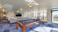 Located at 2184 Fox Valley Center in Aurora, IL, this contemporary amenity lounge is designed to offer a dynamic and social environment for its users. The single-floor space is thoughtfully laid out to accommodate a variety of leisure activities.

At the heart of the room sits a full-size billiard table with a rich wood frame, complemented by a shuffleboard table positioned along a bank of large windows that flood the area with natural light and offer views of the surrounding exterior. Nearby, a high-top counter with bar-style seating lines one wall, decorated with a striking arrangement of botanical wall art panels, providing a casual spot for conversation or casual work.

The lounge area features generous, L-shaped and curved sectional sofas in a warm gray upholstery, arranged around low, round coffee tables, creating an inviting space for relaxation and gathering. A textured brick accent wall serves as a focal point, anchoring a wall-mounted flat-screen television and media console for entertainment.

The space is finished with a combination of tile and carpet flooring, textured ceilings, and globe-style flush-mount light fixtures that add a modern, polished touch. Large windows wrap multiple sides of the room, ensuring the space remains bright and connected to the outdoors throughout the day.