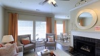This single-floor residence offers an open, flowing layout that seamlessly connects the living and dining areas, creating a cohesive and functional living environment. The living area is anchored by a classic fireplace with a dark tile surround and a white mantel, adding a focal point of warmth and character to the room. A ceiling fan with integrated lighting is centrally positioned, complementing the additional track lighting installed throughout the space.

Large windows dressed with white horizontal blinds and warm orange accent curtains flood the interior with natural light, enhancing the airy, welcoming atmosphere. Crown molding details along the upper walls add a refined architectural touch, while neutral-toned walls provide a versatile backdrop for the space.

The dining area, visible from the living room, features a modern pedestal table paired with upholstered chairs, reinforcing the open-concept design. Hardwood-style flooring in the dining zone transitions smoothly to carpeted areas in the living space. Together, these elements combine to create a bright, comfortable, and well-appointed interior with a warm, inviting ambiance.