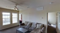 This single-floor residential space offers a functional and straightforward layout designed for comfortable everyday living. The living room serves as the central gathering area, featuring wall-to-wall carpeting that adds warmth underfoot. A ceiling fan with integrated lighting is mounted overhead, providing both air circulation and ambient illumination throughout the space. The room benefits from natural light streaming in through a window, contributing to a bright and welcoming atmosphere. A thermostat is conveniently mounted on the wall, allowing for easy climate control, and HVAC vents are positioned both on the ceiling and upper wall to ensure efficient airflow throughout the home. The neutral, light-toned walls create a clean and versatile backdrop suitable for a variety of interior styles. An open doorway connects the living area to an adjacent bedroom, suggesting a practical and accessible floor plan. The overall ambiance of the space is simple, functional, and move-in ready, making it well-suited for those seeking a low-maintenance residential environment.