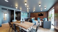This well-appointed single-floor communal space is thoughtfully designed to serve multiple functions within one open, cohesive layout. The room is anchored by a substantial live-edge wood dining table surrounded by sleek black metal chairs, providing an inviting area for group gatherings or casual meals. A woven area rug grounds the dining zone and adds textural contrast to the light hardwood flooring throughout.

Adjacent to the dining area, a comfortable lounge section features plush grey upholstered seating accented with teal throw pillows, arranged around a wall-mounted flat-screen television set against warm, richly toned wood wall paneling. This creates a relaxed, media-friendly retreat within the larger space.

Toward the rear, a compact kitchenette area is outfitted with stainless steel appliances and open shelving displaying small decorative and functional items. A vibrant teal subway tile backsplash serves as a bold design focal point, adding color and personality to the space.

The dark grey ceiling is fitted with an array of globe pendant and flush-mount light fixtures, casting warm, even illumination throughout. The overall aesthetic blends contemporary industrial and mid-century modern influences, resulting in a polished, welcoming environment suited for socializing, dining, working, or unwinding.