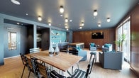 This well-appointed single-floor communal space is thoughtfully designed to serve multiple functions within one open, cohesive layout. The room is anchored by a substantial live-edge wood dining table surrounded by sleek black metal chairs, providing an inviting area for group gatherings or casual meals. A woven area rug grounds the dining zone and adds textural contrast to the light hardwood flooring throughout.

Adjacent to the dining area, a comfortable lounge section features plush grey upholstered seating accented with teal throw pillows, arranged around a wall-mounted flat-screen television set against warm, richly toned wood wall paneling. This creates a relaxed, media-friendly retreat within the larger space.

Toward the rear, a compact kitchenette area is outfitted with stainless steel appliances and open shelving displaying small decorative and functional items. A vibrant teal subway tile backsplash serves as a bold design focal point, adding color and personality to the space.

The dark grey ceiling is fitted with an array of globe pendant and flush-mount light fixtures, casting warm, even illumination throughout. The overall aesthetic blends contemporary industrial and mid-century modern influences, resulting in a polished, welcoming environment suited for socializing, dining, working, or unwinding.