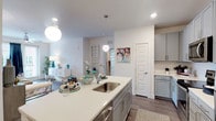 This single-floor residence combines modern design with functional living across a thoughtfully arranged floor plan. The open-concept kitchen serves as the heart of the home, anchored by a generously sized island with a curved edge, quartz countertops, and an undermount sink accented by globe pendant lighting. Gray shaker-style cabinetry lines the walls, complemented by a white subway tile backsplash and a full suite of stainless steel appliances, including a range, microwave, and refrigerator. Wood-look plank flooring flows seamlessly from the kitchen into the living area, where a ceiling fan, recessed lighting, and large windows with sliding glass door access to a private balcony invite natural light throughout. The bedrooms offer plush carpeting, generous window coverage with blinds and curtains, and ceiling fans for added comfort. One bedroom features a walk-in closet and direct access to additional storage. The bathroom is finished with gray cabinetry, a white quartz vanity top, wood-look flooring, and a tub-shower combination. Throughout the residence, crisp white walls and consistent finishes create a cohesive, contemporary aesthetic that feels both inviting and functional.