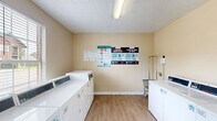 Located at 10624 Glenstone Ct in Baton Rouge, Louisiana, this community laundry facility offers a clean, functional, and well-maintained space designed for convenient everyday use. The room is finished with warm beige walls and durable wood-look hard-surface flooring that is easy to maintain and provides a tidy, welcoming atmosphere. Natural light filters in through multiple windows fitted with horizontal blinds, brightening the interior and creating an open feel. A row of white top-load washing machines and dryers lines one wall, providing ample capacity to serve multiple users. A rolling laundry cart is available for added convenience. The space is equipped with a fire extinguisher mounted on the wall, underscoring a commitment to safety. Informational signage, including laundry tips and usage instructions, is posted on the walls to assist users. The room is accessed through a distinctive red entry door with a glass panel insert, which allows additional natural light into the space. A textured ceiling with overhead lighting ensures the room remains well-lit at all hours. The overall layout is efficient and straightforward, making this laundry facility a practical and accessible amenity for residents at this Baton Rouge location.