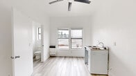 This compact single-bedroom unit is thoughtfully designed to maximize functionality within its modest footprint. The interior is finished throughout with light wood-look luxury vinyl plank flooring and crisp white walls, creating a clean, bright aesthetic that enhances the sense of space.

The main living area serves a dual purpose, incorporating a kitchenette equipped with a compact cooktop, sink, and under-counter refrigerator. A ceiling fan overhead provides air circulation and adds a practical finishing touch. Two side-by-side windows along one wall allow generous natural light to fill the room and offer views of the surrounding rooftop landscape and neighborhood. A wall-mounted heating unit ensures year-round comfort.

The bedroom area is accessed through an interior doorway and shares the same consistent flooring and white wall finish, maintaining a cohesive look throughout. The layout provides a defined separation between the living and sleeping spaces despite the overall compact scale.

The bathroom is neatly appointed with a pedestal sink featuring brushed gold fixtures, a mirrored medicine cabinet for storage, and a walk-in shower with white surround walls. A toilet completes the full bathroom. The all-white finish keeps the space feeling clean and open.

Overall, this unit delivers a minimalist, move-in-ready living environment with efficient use of every square foot.