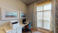 Located at 557 Roanoke Rd in Daleville, Virginia, this single-floor space is designed with both functionality and comfort in mind. The interior showcases a cohesive aesthetic defined by warm gray walls, crisp white baseboards, and light hardwood-style flooring that runs throughout.

The centerpiece of the space is a dedicated business center area, outfitted with built-in cabinetry featuring white shaker-style cabinet doors and stone-look granite countertops that provide generous workspace. The workstation is well-equipped for productivity, with ample surface area for computing and printing needs. Framed artwork adorns the walls above the desk, adding a polished, professional touch to the environment.

Natural light is a defining feature of the space, with large windows dressed in wood blinds and decorative patterned drip curtains in a multicolor geometric print. The windows allow abundant daylight to filter in, creating a bright and welcoming atmosphere. A glass-paneled interior door with a white frame connects to an adjacent area, reinforcing the open and airy feel of the layout.

The space also includes an open flex room with clean, neutral walls and the same warm flooring, offering versatility for various uses. Overall, the space presents a clean, modern, and well-maintained environment suited for professional or community-oriented purposes.