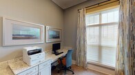 Located at 557 Roanoke Rd in Daleville, Virginia, this single-floor space is designed with both functionality and comfort in mind. The interior showcases a cohesive aesthetic defined by warm gray walls, crisp white baseboards, and light hardwood-style flooring that runs throughout.

The centerpiece of the space is a dedicated business center area, outfitted with built-in cabinetry featuring white shaker-style cabinet doors and stone-look granite countertops that provide generous workspace. The workstation is well-equipped for productivity, with ample surface area for computing and printing needs. Framed artwork adorns the walls above the desk, adding a polished, professional touch to the environment.

Natural light is a defining feature of the space, with large windows dressed in wood blinds and decorative patterned drip curtains in a multicolor geometric print. The windows allow abundant daylight to filter in, creating a bright and welcoming atmosphere. A glass-paneled interior door with a white frame connects to an adjacent area, reinforcing the open and airy feel of the layout.

The space also includes an open flex room with clean, neutral walls and the same warm flooring, offering versatility for various uses. Overall, the space presents a clean, modern, and well-maintained environment suited for professional or community-oriented purposes.
