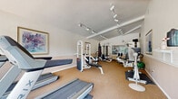 This well-equipped fitness center in Denver, Colorado offers a functional and inviting workout environment within a single, efficiently designed exercise room. The space features vaulted ceilings that lend an open, airy feel, complemented by track lighting that ensures bright, even illumination throughout. Large windows along one wall flood the room with natural light and provide pleasant outdoor views of surrounding greenery, enhancing the overall workout experience.

The flooring consists of durable, cushioned carpet-style material well-suited for fitness use, offering comfort underfoot during exercise. The room is outfitted with a comprehensive selection of equipment, including multiple treadmills, a stationary bike, a multi-station cable weight machine, a captain's chair for core training, a punching bag, resistance bands, a stability ball, and a flat bench. A wall-mounted television adds a convenient entertainment option for users during cardio sessions.

Mirrors are positioned strategically to assist with form and to visually expand the space. The neutral, two-tone wall treatment — white upper walls with a warm beige lower accent — creates a clean and focused atmosphere. A glass-paneled door provides easy access and connects the fitness room to the broader community. Overall, the space delivers a practical and complete fitness facility designed to support a wide range of exercise routines.