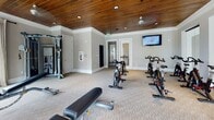 This fitness center offers a thoughtfully designed workout environment that balances functionality with refined aesthetics. The space is immediately distinguished by its rich, warm wood-planked ceiling, which is complemented by recessed lighting and a ceiling fan, creating an inviting and comfortable atmosphere for exercise. Large, floor-to-ceiling windows dressed with sheer linen curtains flood the interior with natural light and offer views of surrounding greenery and a nearby pool area, making workouts feel open and energizing. The flooring throughout is covered in a neutral, textured carpet that adds softness underfoot. The fitness area is well-equipped, featuring a multi-station cable machine, an adjustable weight bench, a full rack of dumbbells, and a row of cycling bikes accompanied by a wall-mounted television — ideal for group or solo cardio sessions. A full-length mirror along one wall enhances the sense of space and supports form-focused training. Glass panel partitions offer a transparent view into adjacent areas, adding an architectural openness to the layout. Additional amenities include a fire extinguisher station and access to a restroom. The overall ambiance is polished yet welcoming, designed to support a comprehensive fitness routine in a bright, well-maintained setting.
