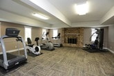 This fitness space is thoughtfully designed to support a well-rounded workout routine within a comfortable, functional environment. The room features textured, multi-tone carpet tiles throughout the exercise area, providing cushioning underfoot and a clean, organized aesthetic. Neutral taupe-toned walls with crisp white trim create a calm, focused atmosphere.

The cardio section is equipped with a treadmill, elliptical trainer, and a recumbent-style cycle, each positioned to take advantage of natural light streaming through multiple windows fitted with white blinds. A standout architectural feature is the full brick fireplace, which anchors one wall and adds warmth and character rarely found in a fitness setting.

A multi-station strength training machine and stability ball cater to resistance and flexibility training, while a wall-mounted flat-screen television provides entertainment during workouts. Overhead fluorescent lighting ensures consistent illumination throughout the space.

Adjacent to the fitness area, a lounge space with tile flooring and bistro-style tables and chairs offers a convenient spot to rest or socialize. A refrigerator is also visible in this area, adding practical convenience for residents or users of the facility.