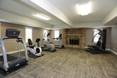 This fitness space is thoughtfully designed to support a well-rounded workout routine within a comfortable, functional environment. The room features textured, multi-tone carpet tiles throughout the exercise area, providing cushioning underfoot and a clean, organized aesthetic. Neutral taupe-toned walls with crisp white trim create a calm, focused atmosphere.

The cardio section is equipped with a treadmill, elliptical trainer, and a recumbent-style cycle, each positioned to take advantage of natural light streaming through multiple windows fitted with white blinds. A standout architectural feature is the full brick fireplace, which anchors one wall and adds warmth and character rarely found in a fitness setting.

A multi-station strength training machine and stability ball cater to resistance and flexibility training, while a wall-mounted flat-screen television provides entertainment during workouts. Overhead fluorescent lighting ensures consistent illumination throughout the space.

Adjacent to the fitness area, a lounge space with tile flooring and bistro-style tables and chairs offers a convenient spot to rest or socialize. A refrigerator is also visible in this area, adding practical convenience for residents or users of the facility.