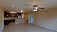 This single-story space showcases an open-concept design that seamlessly connects the living area and kitchen. The living space is finished with large, light-toned ceramic tile flooring that extends throughout, contributing to a clean and cohesive aesthetic. A ceiling fan with an integrated light fixture is centrally mounted, providing both air circulation and ambient lighting.

The kitchen is defined by dark wood cabinetry that provides a warm contrast to the neutral, beige-toned walls. A raised breakfast bar acts as a natural divider between the kitchen and living area, offering casual seating and additional counter space. Pendant lights hang above the bar, complementing the flush-mount ceiling fixture overhead. A white refrigerator anchors the kitchen, which benefits from its own dedicated lighting.

Doorways visible from the main living area suggest access to additional rooms, adding to the overall functionality of the layout. The warm wall tones and consistent flooring create a unified, welcoming ambiance throughout the space. Overall, the open floor plan and practical finishes make this a functional and comfortable single-floor residence.