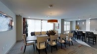 This well-appointed single-floor space presents a polished and professional atmosphere, ideal for dining or group gatherings. The centerpiece of the room is a large dark-wood dining table surrounded by stylish cream upholstered chairs with nailhead trim detailing, offering comfortable seating for a sizable group. A bold orange drum pendant light hangs overhead, adding a warm focal point and a touch of contemporary flair to the neutral palette.

Floor-to-ceiling windows dressed with sheer vertical blinds line one wall, flooding the interior with abundant natural light while maintaining a sense of privacy. Additional windows with plantation shutters on an adjacent wall further enhance the bright and airy ambiance. The light-toned walls are accented by rich red architectural panels, adding depth and visual interest to the space.

Patterned carpeting in muted tones grounds the room and adds texture underfoot. A decorative floral centerpiece adorns the dining table, while tall indoor plants in the corners bring a touch of greenery and life to the setting. A framed piece of artwork on the wall contributes to the curated, refined aesthetic. The overall design strikes a balance between elegance and functionality, creating an inviting environment suited for both formal and casual occasions.