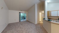 This single-floor, 3-bedroom, 2-bathroom home offers a well-organized layout designed for comfortable everyday living. The living area is highlighted by striking vaulted ceilings that create an open, airy atmosphere, with soft neutral carpeting underfoot and a pendant light fixture adding a warm focal point. A sliding glass door leads to a private balcony overlooking lush greenery, bringing natural light deep into the space. The kitchen is efficiently designed in a U-shaped configuration, featuring white cabinetry with warm wood trim accents, a full suite of appliances including a refrigerator, electric range, microwave, and dishwasher, and a pass-through counter that connects to the dining and living areas. Each of the three bedrooms is carpeted and features windows with blinds that allow natural light while maintaining privacy. The bedrooms offer clean, neutral walls and ample space for furnishings. Both bathrooms share a similar design, each featuring a single vanity with a wide mirror, wood-toned cabinetry, and a shower-tub combination. The bathrooms are finished with hardwood-style flooring, providing a practical and clean contrast to the carpeted living spaces. Overall, the home presents a clean, functional aesthetic with a neutral palette throughout.