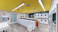 This well-appointed laundry facility offers a clean, modern environment designed to make laundry day as efficient and pleasant as possible. The single-floor space is immediately eye-catching, featuring a bold yellow ceiling accented by bright linear lighting that floods the room with a warm, energetic glow. The walls are kept crisp white, with a teal accent wall providing a striking visual contrast that adds personality to the space. Durable wood-look flooring runs throughout, lending a polished yet practical finish underfoot. A long, built-in countertop with a light wood surface runs centrally through the room, providing ample space for sorting and folding laundry. Rows of top-load washing machines are neatly lined up along this counter, while a generous bank of stacked front-load dryers lines the adjacent wall, offering substantial capacity to accommodate high demand. Small potted plants are thoughtfully placed along the counter, adding a touch of greenery and warmth to the utilitarian setting. The space also includes practical amenities such as laundry baskets and a stainless steel waste bin. Ventilation is handled by ceiling-integrated HVAC vents, ensuring the environment remains comfortable throughout. Overall, the facility strikes an effective balance between function and thoughtful design.