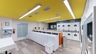 This well-appointed laundry facility offers a clean, modern environment designed to make laundry day as efficient and pleasant as possible. The single-floor space is immediately eye-catching, featuring a bold yellow ceiling accented by bright linear lighting that floods the room with a warm, energetic glow. The walls are kept crisp white, with a teal accent wall providing a striking visual contrast that adds personality to the space. Durable wood-look flooring runs throughout, lending a polished yet practical finish underfoot. A long, built-in countertop with a light wood surface runs centrally through the room, providing ample space for sorting and folding laundry. Rows of top-load washing machines are neatly lined up along this counter, while a generous bank of stacked front-load dryers lines the adjacent wall, offering substantial capacity to accommodate high demand. Small potted plants are thoughtfully placed along the counter, adding a touch of greenery and warmth to the utilitarian setting. The space also includes practical amenities such as laundry baskets and a stainless steel waste bin. Ventilation is handled by ceiling-integrated HVAC vents, ensuring the environment remains comfortable throughout. Overall, the facility strikes an effective balance between function and thoughtful design.
