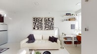 This thoughtfully designed single-floor space embraces a clean, modern aesthetic built around a crisp white palette throughout. The open living area serves as the heart of the home, anchored by a cream upholstered sofa accented with dark throw pillows and a cozy knit blanket. A striking pair of bold black-and-white abstract art pieces commands attention on the wall above, complemented by floating white shelves displaying curated décor. A transparent acrylic coffee table and a cowhide area rug add texture and visual interest to the space. Natural light pours in through a glass-paneled exterior door, which opens to an outdoor view and enhances the airy, open feel of the living area. A dedicated media wall features a warm wood-toned dresser beneath a flat-screen TV, flanked by open shelving for additional storage. An adjacent dining nook with warm-toned upholstered chairs provides a defined space for meals. The compact kitchen is efficiently laid out with rich dark-stained cabinetry, grey countertops, stainless steel appliances, and a deep undermount sink. The bathroom continues the refined aesthetic with marble-look wall tile, a white vanity, and clean, minimal finishes throughout. The space is cohesive, functional, and thoughtfully appointed.