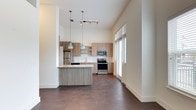 This 1-bedroom, 1-bathroom unit offers a thoughtfully designed, open-concept floor plan that maximizes functionality and flow. The living and kitchen areas share a seamlessly connected layout, anchored by rich, dark hardwood flooring that extends throughout the space.

The contemporary kitchen is a standout feature, equipped with stainless steel appliances including a refrigerator, microwave, and range. Warm-toned wood cabinetry with open shelving provides ample storage, while a peninsula with a light stone countertop serves as both a workspace and a casual dining or entertaining area. A subway tile backsplash adds a clean, modern touch. The kitchen is illuminated by stylish pendant lights that hang at varying heights, enhancing the sense of vertical space created by the notably tall ceilings.

Large windows along the living area allow an abundance of natural light to fill the space, contributing to an open and airy ambiance. Track lighting supplements illumination throughout the main living zone.

The overall aesthetic is modern and clean, combining neutral tones, natural wood textures, and contemporary fixtures to create a cohesive and inviting environment. The single-floor layout ensures easy accessibility and a practical living experience throughout the unit.