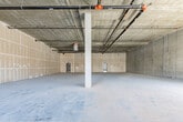 This single-floor commercial shell space presents a blank canvas ideal for a wide range of retail, restaurant, or studio uses. The open floor plan is expansive and column-supported, with structural concrete columns positioned throughout that lend an industrial character while defining the spatial layout. Underfoot, polished concrete floors extend across the entire space, providing a durable and versatile foundation suited to virtually any build-out concept.

The exposed concrete ceiling reveals the building's raw structural framework, with visible conduit runs, piping, and mechanical infrastructure overhead — a hallmark of modern industrial-style architecture that adds visual depth and flexibility for future lighting and system integration. Drywall has been installed and taped along interior walls, indicating the space is in an active shell-condition phase, ready for finish work.

Toward the street-facing facade, large multi-pane windows and glass-paneled entry doors flood the front portion of the space with natural light and provide prominent visibility to the surrounding streetscape. Multiple entry points are accessible from the exterior, enhancing accessibility and foot traffic potential. Interior access doors along the rear walls suggest additional utility or egress connections. The overall ambiance is clean, open, and industrial — a highly functional framework awaiting customization.