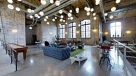 This captivating single-floor space masterfully combines industrial heritage with contemporary design. The defining architectural feature is the expansive exposed brick wall, aged to a warm patina, which stretches upward to meet the dramatically high ceilings lined with visible steel beams and ductwork — hallmarks of authentic industrial conversion. Flooding the interior with natural light are a series of tall, arched multi-pane windows framed in dark metal, lending both grandeur and character to the space. Suspended from the ceiling is a stunning installation of spherical globe pendant lights at varying heights, creating an ambient, gallery-like atmosphere throughout the open floor plan. The polished concrete floors reflect light beautifully, enhancing the sense of openness and scale. The interior is furnished with a mix of eclectic and creative pieces, including a deeply tufted velvet sofa, accent seating, and casual gathering areas that define zones within the open layout. A green turf rug adds a playful, unexpected contrast to the industrial backdrop. The overall ambiance is one of creative energy — raw yet refined, open yet intimate — making it a versatile and visually compelling space suited for a wide range of uses.
