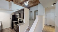 This distinctive loft-style space blends industrial character with modern updates, highlighted by dramatic exposed wooden beam ceilings that run throughout the unit and lend a warm, rustic ambiance. Soaring ceiling heights in the main living area create an open, airy atmosphere, with the structural beams serving as a standout architectural feature.

The kitchen is well-appointed, featuring dark espresso-toned cabinetry, granite-look countertops, and a full suite of stainless steel appliances including an electric range, over-the-range microwave, and refrigerator. Wood-look flooring in the kitchen adds to the space's contemporary feel, while a ceiling fan with a pendant light overhead provides both function and style.

The living and bedroom areas are finished with plush carpet and benefit from the dramatic height of the exposed beam ceilings. A large multi-pane window in the bedroom invites natural light and frames views of the surrounding brick architecture, reinforcing the building's historic character. Painted brick walls in the bedroom further emphasize the loft aesthetic.

The bathroom offers a clean, straightforward layout with a combination tub and shower surrounded by classic white tile, a toilet, and a vanity with a mirror. A second bathroom space provides an additional shower option. Throughout, the space strikes a balance between preserved industrial character and practical, updated living features.
