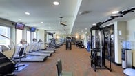 This fitness center offers a comprehensive workout environment designed to accommodate a wide variety of exercise routines. The space is laid out on a single floor, with an open, elongated floor plan that creates a sense of generous room to move freely between stations. A row of treadmills lines one side of the facility, providing ample cardio options, while an assortment of ellipticals, stationary bikes, and other aerobic machines fill the middle section. On the opposite side, a selection of cable machines and strength training equipment offers a full range of resistance workouts. The flooring features a patterned carpet in warm tones, adding color and comfort underfoot. Overhead, recessed lighting is evenly distributed throughout the ceiling, ensuring bright, consistent illumination across the entire space. Ceiling fans are mounted at intervals, promoting air circulation during intense workouts. Wall-mounted televisions are positioned for easy viewing from the cardio equipment. Large mirrors along one wall visually expand the space and allow users to monitor their form. The overall ambiance is practical and energizing, designed to support an effective and comfortable fitness experience.