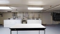 This single-floor laundry facility offers a practical and utilitarian space designed to accommodate shared use. The room features exposed concrete block walls painted white, visible overhead piping, and bright fluorescent panel lighting that ensures strong, even illumination throughout. Several top-load washing machines and matching dryers are arranged along the back wall, providing ample capacity for multiple users. A water heater is also present, supporting the room's plumbing infrastructure. A large central folding table with a white surface and dark trim edge is positioned in the foreground, offering a generous workspace for sorting and folding laundry. Informational notices are posted on the wall, indicating organized building management. The overall aesthetic is straightforward and functional, prioritizing utility over decoration. The exposed mechanical elements, including ductwork and piping along the ceiling and walls, reflect the room's practical, no-frills character. The space is clean and well-lit, making it a convenient and efficient shared laundry resource.